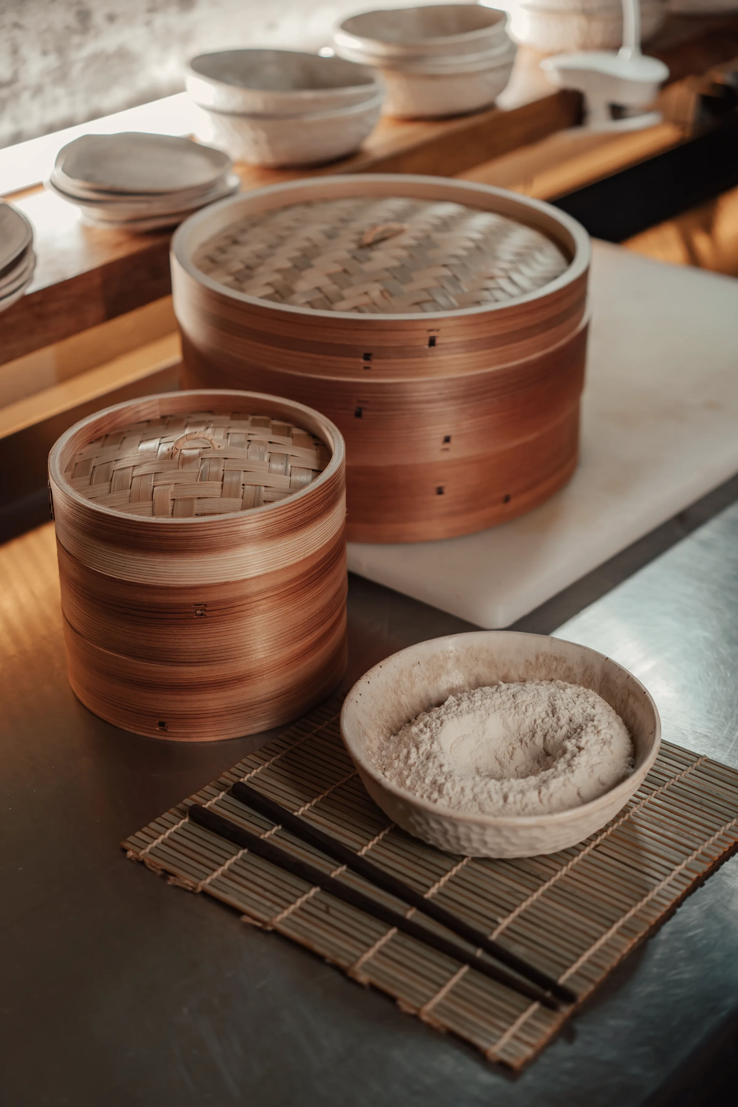 Stacked bamboo steamer baskets beside a bowl of flour on a wooden bench