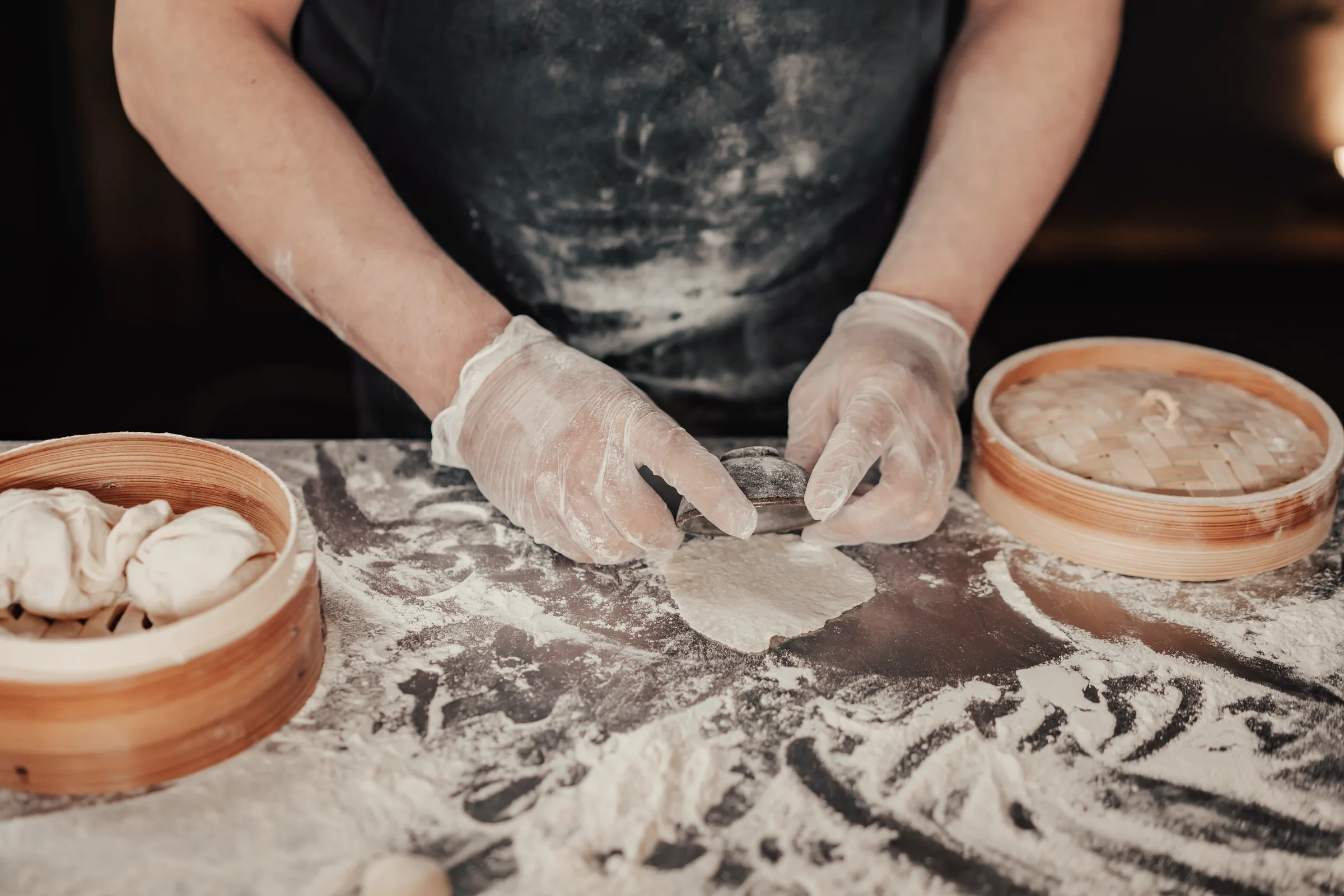 Hands flattening a dumpling wrapper on a floured board beside bamboo steamers